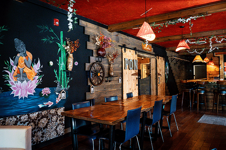 Rustic restaurant interior with wooden tables, Buddha mural, hanging lights, and floral decor.