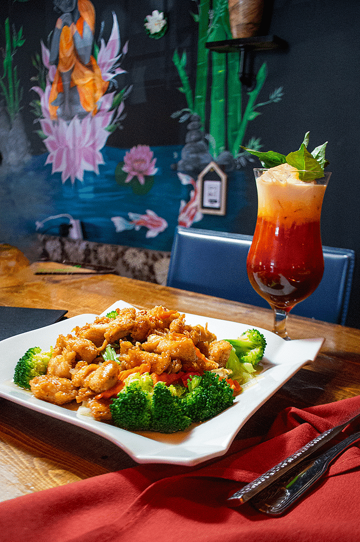 A plate of stir-fried chicken with broccoli and tropical cocktail on a wooden table near a mural.
